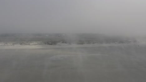 Drohne-Fliegt-über-Sehr-Nebligen-Strand-Entlang-Des-Windes-Und-Des-Aufgewirbelten-Sandes-In-Richtung-Dünen