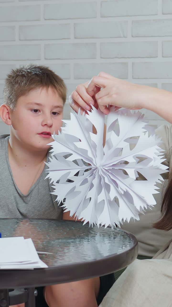 white boy watches paper snowflake unfold, hands revealing intricate cut pattern on round decoration closeup of curious child apprentice learning craft at glass table while mother guides as teacher