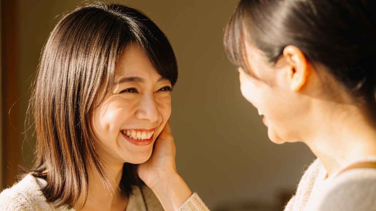 右側の女性が左頬に手を当て、笑いながら家で優しく顔を包み込んでいる。ニットを着ている。