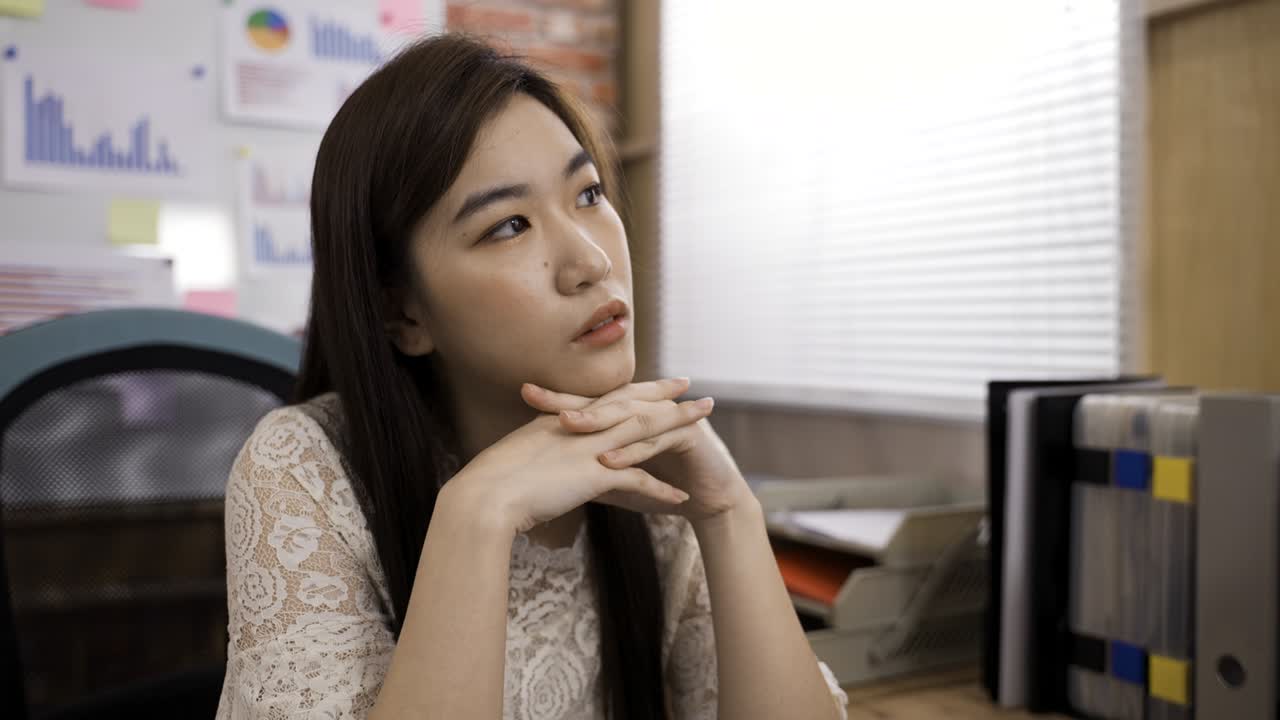ภาพถ่ายครึ่งตัวของผู้ประกอบการหญิงชาวเกาหลีกำลังวางมือใต้คาง จ้องมองออกไปไกลๆ และพึมพำกับตัวเองในที่ทำงานที่สว่างสดใส