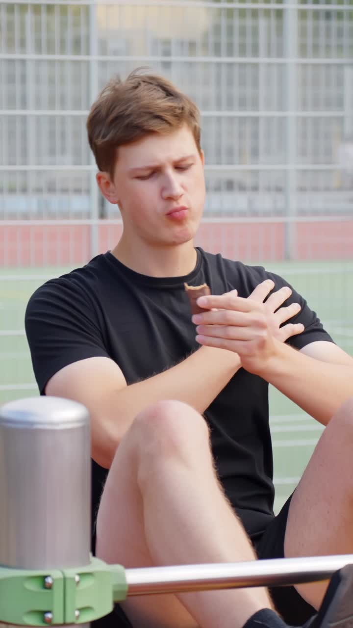 Fit young man does crunches and bites into chocolate bar as reward after exercise outdoors