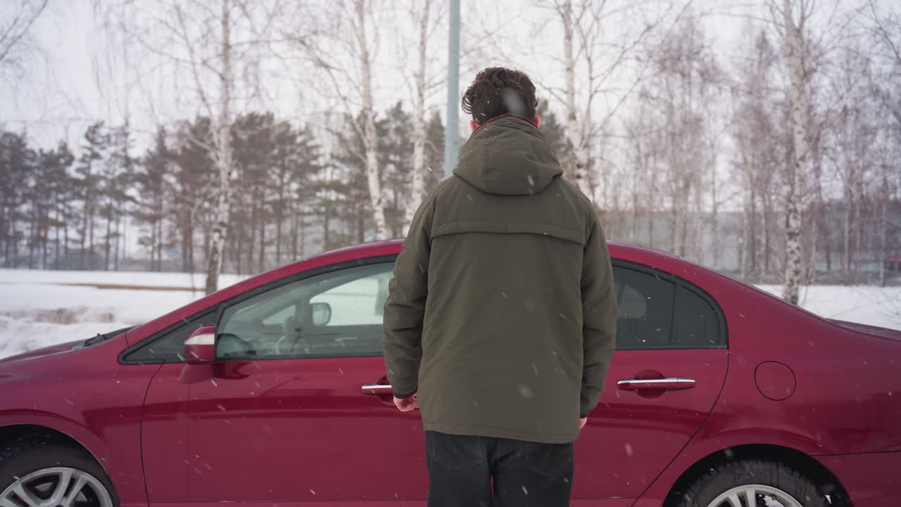 Back view of boy wearing winter jacket walking across snow covered ground toward car door before entering vehicle during cold cloudy winter day in urban parking lot with parked cars and buildings
