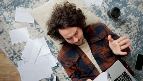 a confident brunette guy with curly hair in a checkered shirt makes notes in his notebook and then tears out a sheet from it and does it again while lying on the floor on a pillow around sheets of paper and a laptop