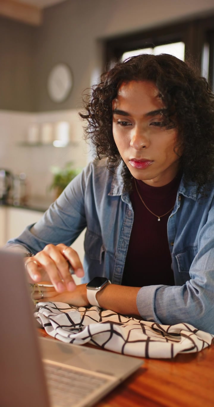 Vertical video: Non-binary person glancing then scrolling laptop and jotting work notes at kitchen