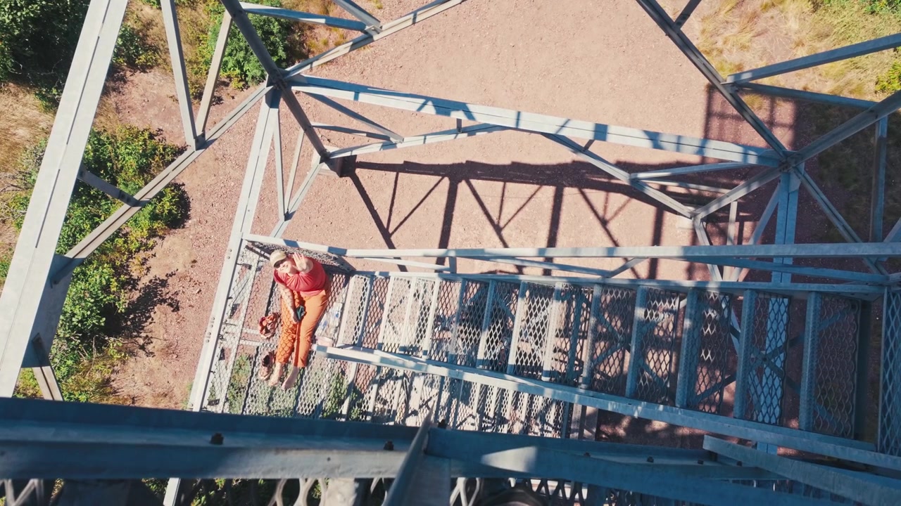 A woman sitting on a metal staircase of a watchtower waving up at a downward shot with a dirt road background on a sunny day.