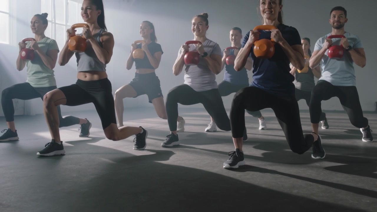 Responding to coach cue, class lifting kettlebells and lunging at gym, tank tops for workout