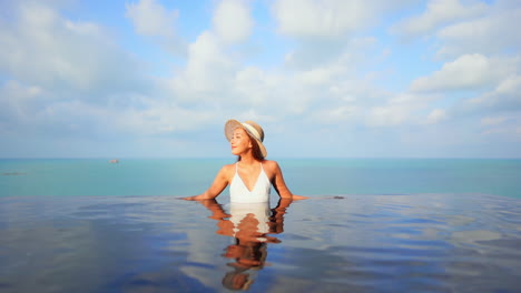 Junge-Asiatische-Frau-Mit-Hut,-Die-Sich-Im-Infinity-Pool-Mit-Horizont-Im-Hintergrund-Entspannt