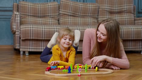 Mother-with-little-daughter-child-girl-riding-toy-train-on-wooden-railway-blocks-board-game-at-home