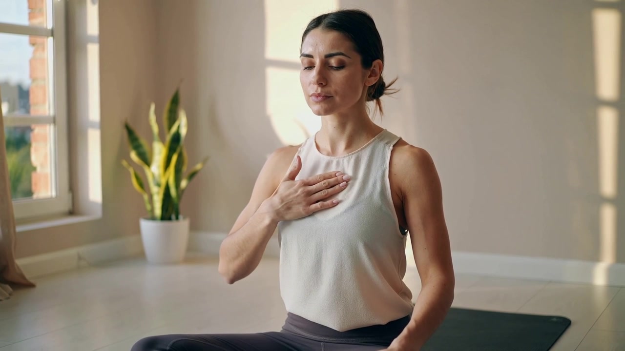 Žena v legínách a tílku s rukou na hrudi praktikující meditaci u okna s podložkou a rostlinou.
