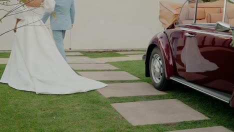 bride in white gown and groom walking past a vintage car on a garden path