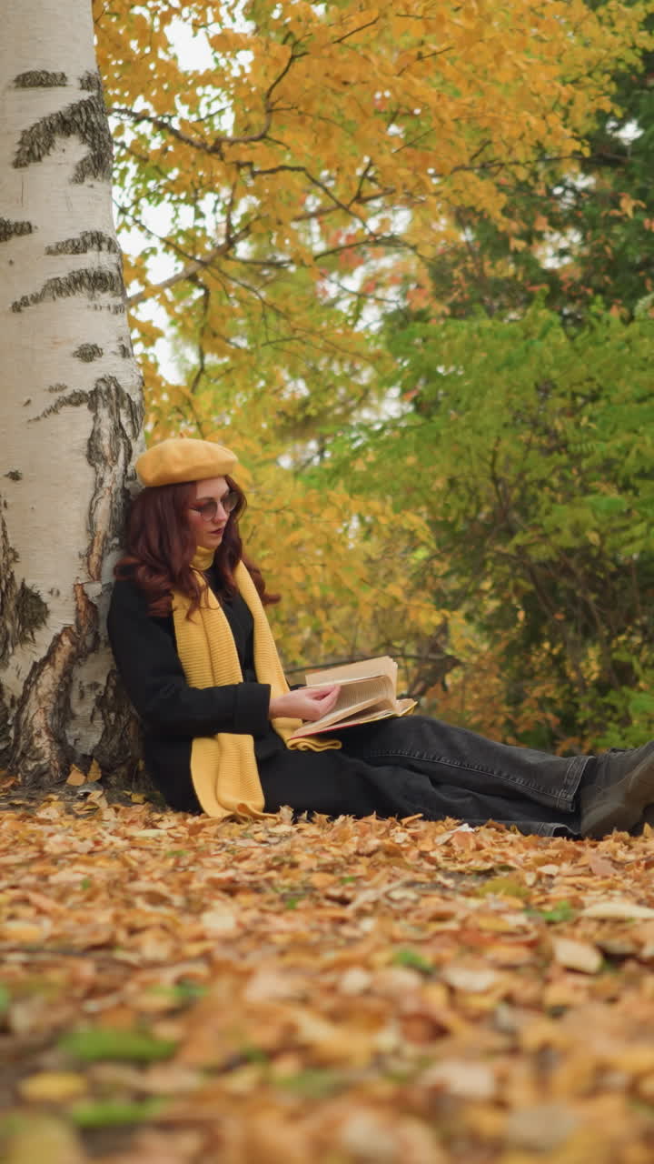 una elegante dama con boina amarilla se relaja al aire libre, apoyándose en un árbol mientras hojea libros, rodeada de hojas doradas de otoño esparcidas por el suelo, disfrutando de la tranquila atmósfera estacional