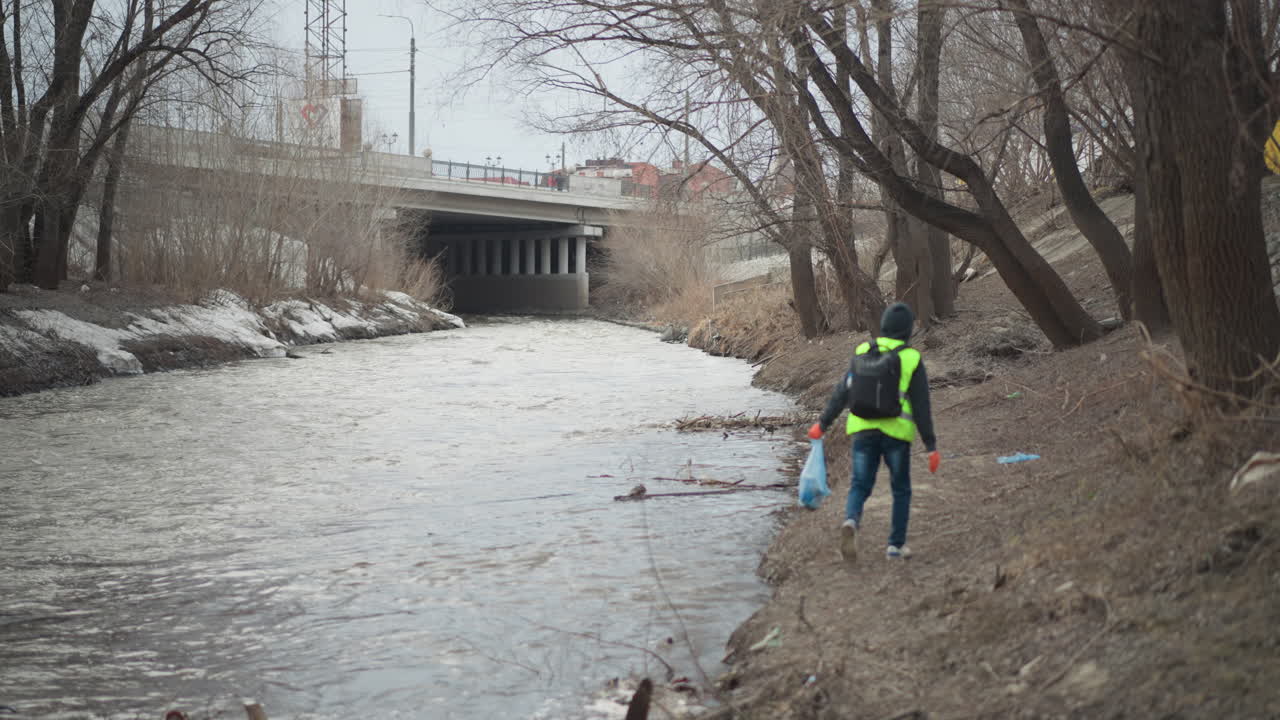 Volunteer in reflective vest walks along polluted riverbank carrying blue bag filled with collected litter during environmental cleanup effort to protect water, restore nature