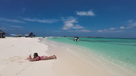 woman lying face down on white sand beach taking photos with phone, azure caribbean sea background