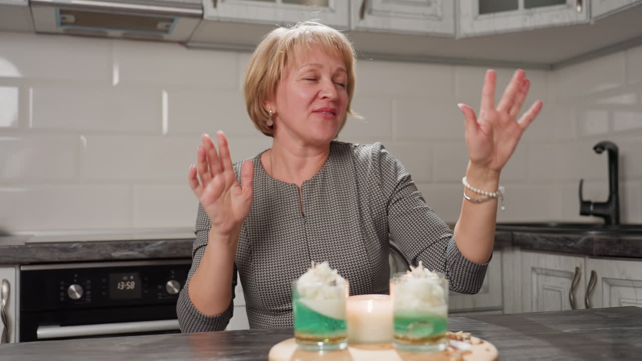 Beautiful grandma seated in kitchen joyfully sings and grooves to music with expressive hand movements and bright smile, radiating happiness and energy while enjoying melody in relaxed home setting