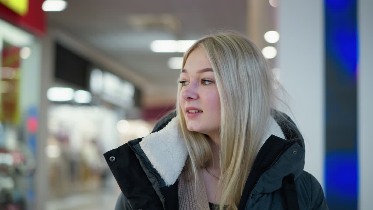 미소 짓고 쇼핑을 마친 여인은 잘 조명된 쇼핑몰을 걸어가며 배경에 소매점과 장식적인 박이는 불빛으로 분위기를 향상시키는 사려 깊게 주위를 둘러보고 있습니다.