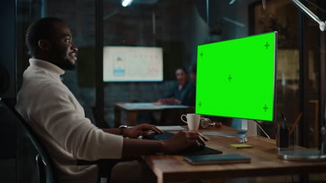 un apuesto especialista afroamericano negro que trabaja en una computadora de escritorio con una pantalla verde en una oficina creativa ocupada. el gerente masculino lleva un suéter de cuello de tortuga blanco casual.
