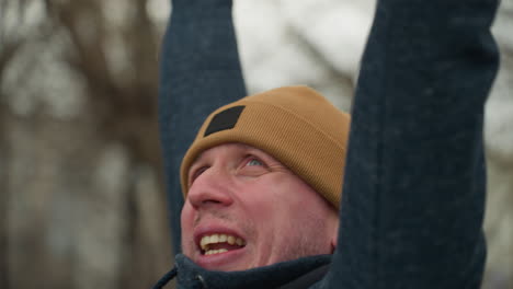 close-up of a coach working out on red iron bars, visibly tired as he moves across the iron bar drops down, the blurred background features bare trees