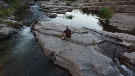 Mann-Sitzt-Auf-Einem-Felsen-Mitten-Im-Fluss-Und-Ruht-Sich-Aus,-Während-Er-Die-Aussicht-Genießt