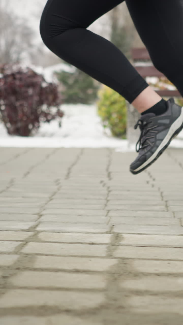 close up leg view of lady jogging in black sneakers and leggings along snowy park pathway with frosty winter trees bushes distant residence visible breath conveying cold fitness motion