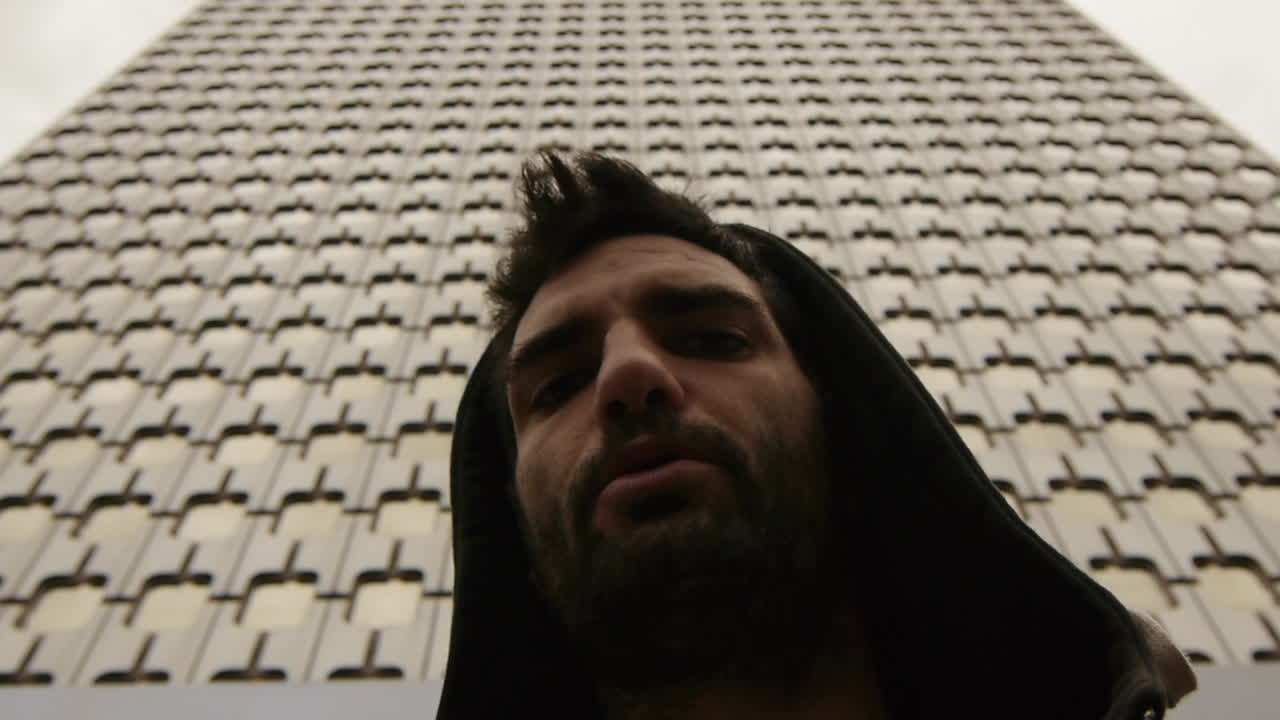 Moody close-up of a man in a hoodie against a geometric building facade, evoking urban isolation and raw introspection