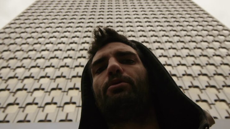 Primo piano intenso di un uomo con una felpa con cappuccio sullo sfondo di una facciata geometrica, che evoca isolamento urbano e cruda introspezione