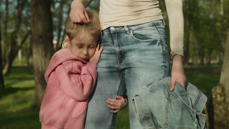 sleepy little girl leans on mother thigh standing in park