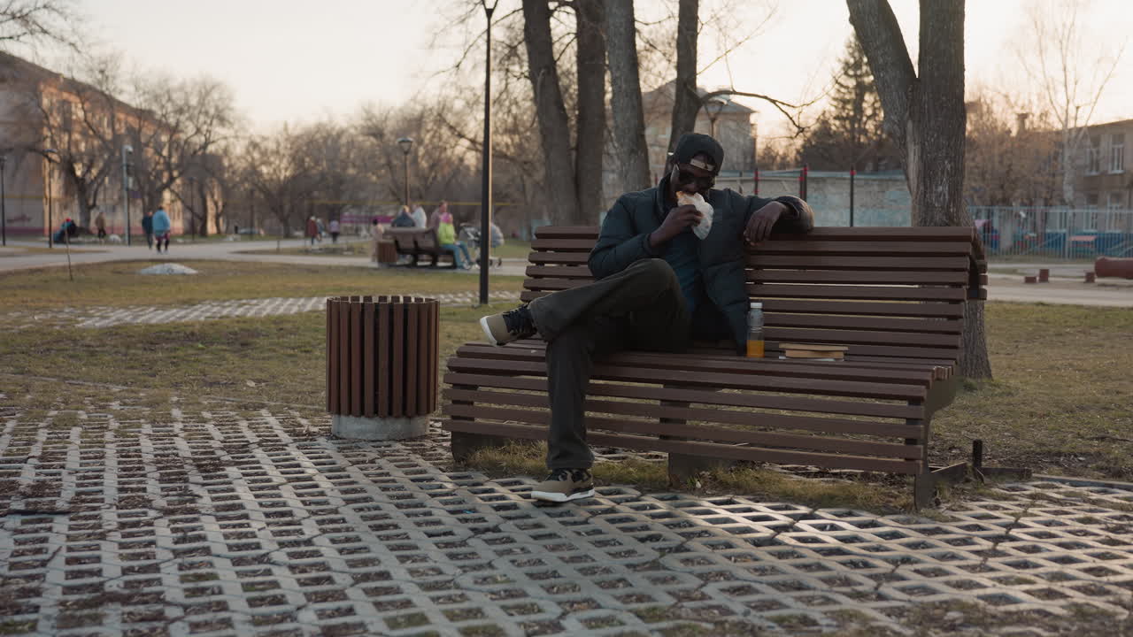 Dark skin man sitting on park bench eating snack with bottled drink beside him, wearing cap and sunglasses, relaxed posture in outdoor park during sunset with people in background