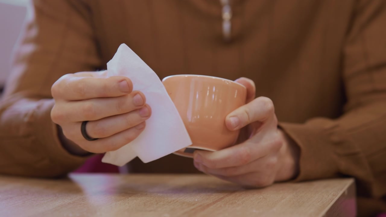 Primo piano di mani che tengono una tazza di ceramica e la puliscono con un tovagliolo di carta. La persona indossa una maglietta a maniche lunghe marrone, visibile dal busto in giù, per mostrare un'azione di pulizia accurata. Illuminazione interna soffusa.