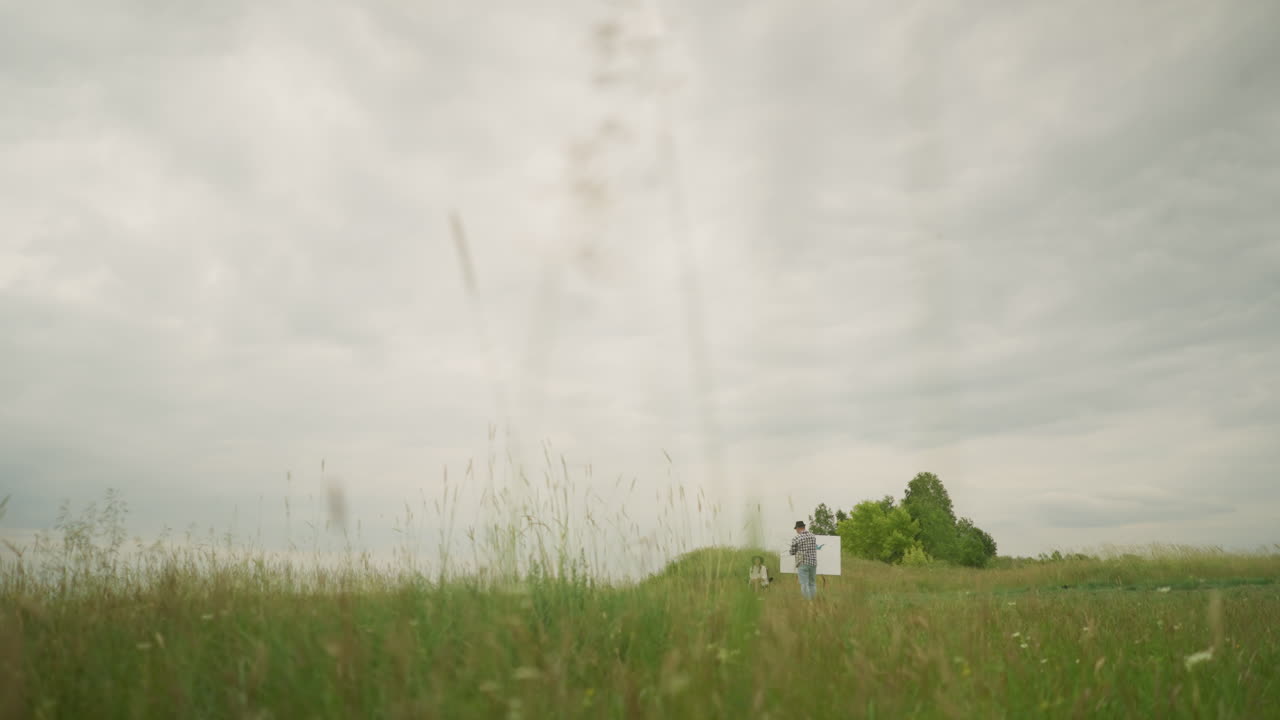 A craftsman wearing a hat and checkered shirt is intently focused on painting on a board in the middle of a lush grass field under a cloudy sky. Nearby, a woman sits comfortably on a chair