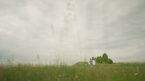 a craftsman wearing a hat and checkered shirt is intently focused on painting on a board in the middle of a lush grass field under a cloudy sky. nearby, a woman sits comfortably on a chair