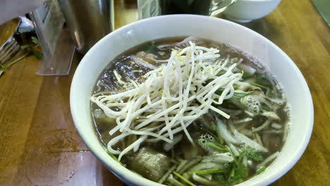 fresh vietnamese noodle soup on a table in a bowl adding ingredients, handheld shot