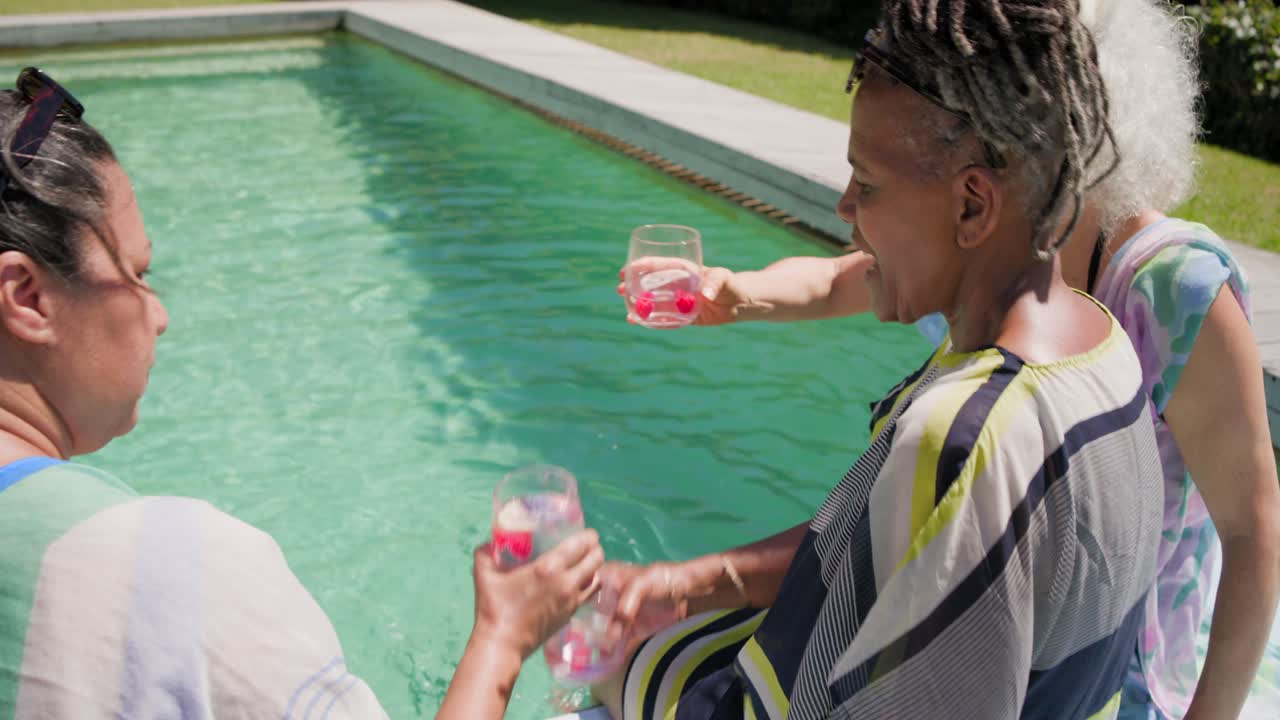 Amigas diversas passando copos sem haste, brindando com bebidas cor-de-rosa e saboreando-as na piscina do quintal