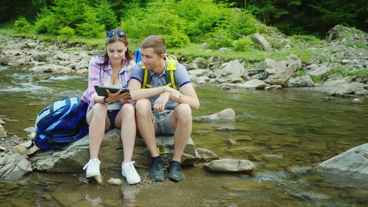 una pareja joven de turistas disfruta de una tableta en un viaje sentado en una roca cerca de un río de montaña 4k video