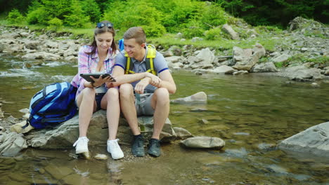 una pareja joven de turistas disfruta de una tableta en un viaje sentado en una roca cerca de un río de montaña 4k video