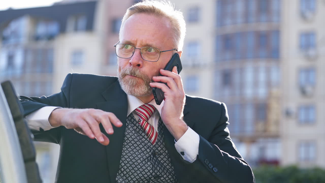 Close-up portrait of mature businessman with beard and eyeglasses. Senior man in suit sitting outdoors and talking on phone gesticulating a lot.