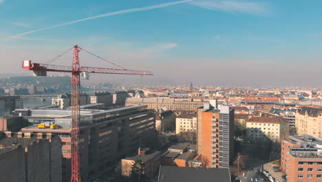 ascending aerial shot over a city with a red crane working on construction and turns around, showing the top of the buildings and a cityscape