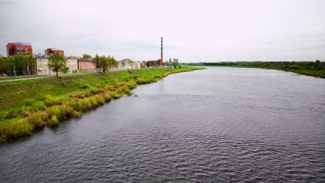 Wide view of calm Daugava river bordered by buildings and greenery, featuring a tall chimney in the distance. Shot at Daugavpils, Latgale, Latvia