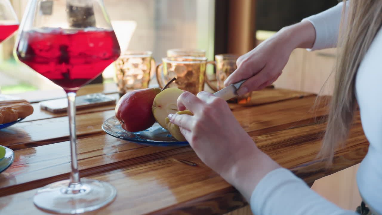 Woman with long brown hair slices apple with knife on wooden table inside sunlit kitchen, red iced drink in glass sits nearby, casual setting captures peaceful moment of simple snack preparation