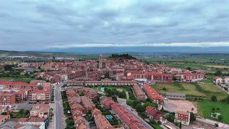 Smooth aerial orbit around Navarrete, La Rioja. Scenic view of historic town, church tower, red rooftops, vineyards and green countryside under cloudy sky