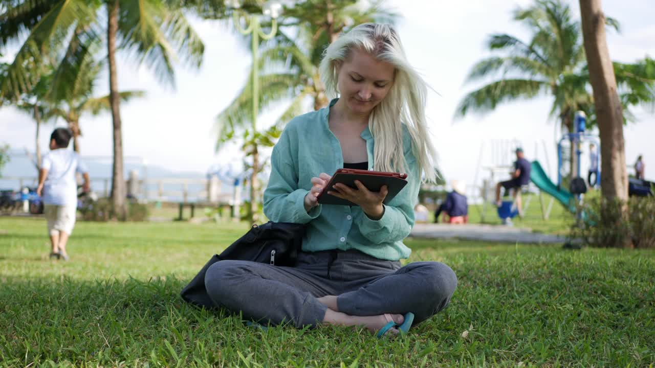 Beautiful slim woman with long blonde hair in green shirt sits on the ground and using smartphone over background the park. Girl on the square touching screen and smile