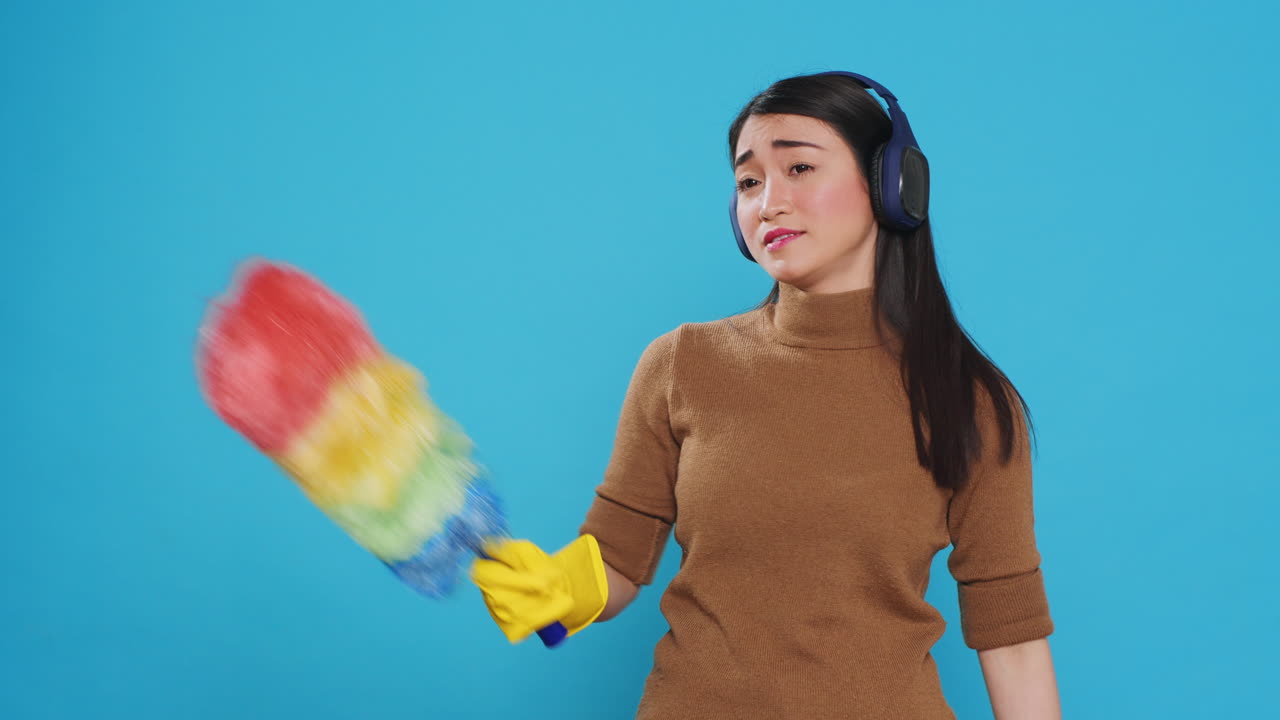 Exhausted sleepy housekeeper wearing headphones while doing cleaning in house using feather duster