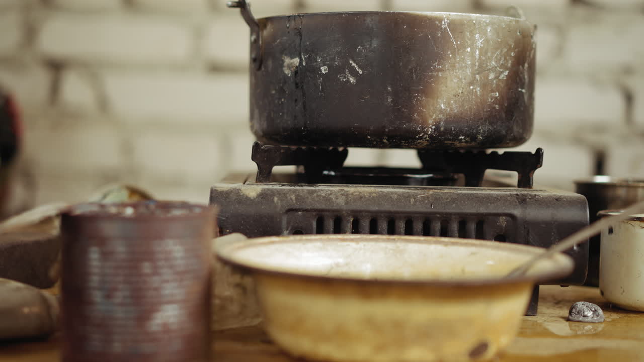Old soot covered pot sits atop rusty gas burner with surrounding stained plates, corroded tin can, and grimy cooking tools, suggesting hardship, poverty, or outdoor survival in harsh environment