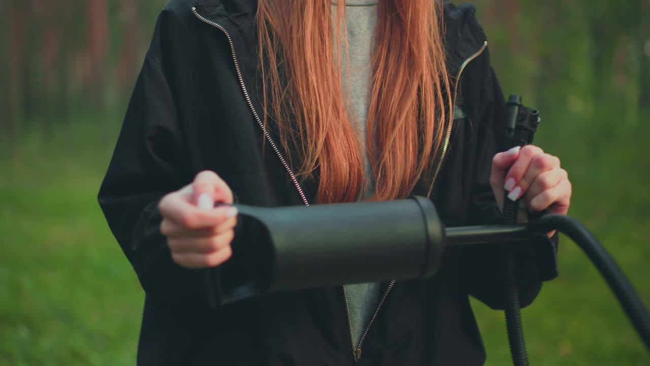 Close up of woman with long reddish hair wearing black jacket and grey shirt while holding and pumping nozzle with both hands in forest setting