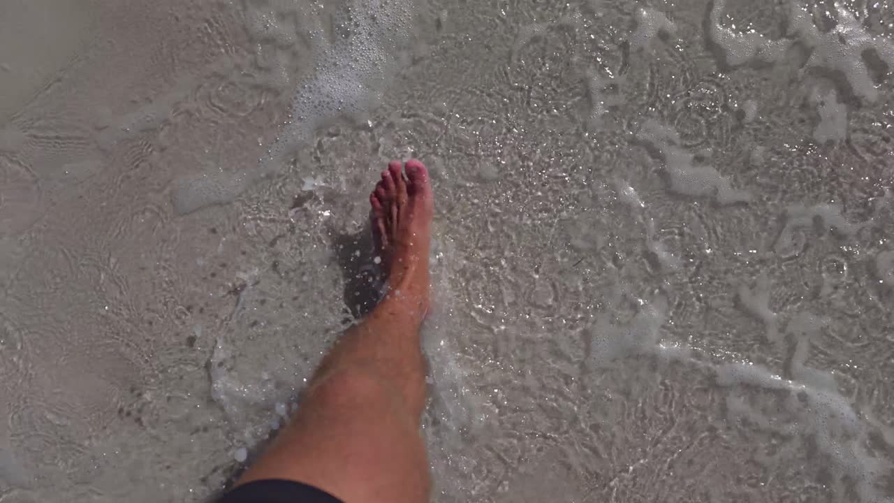 A barefoot person steps into shallow ocean waves as gentle water flows over smooth sand. The slow-motion captures each splash and bubble in detail.