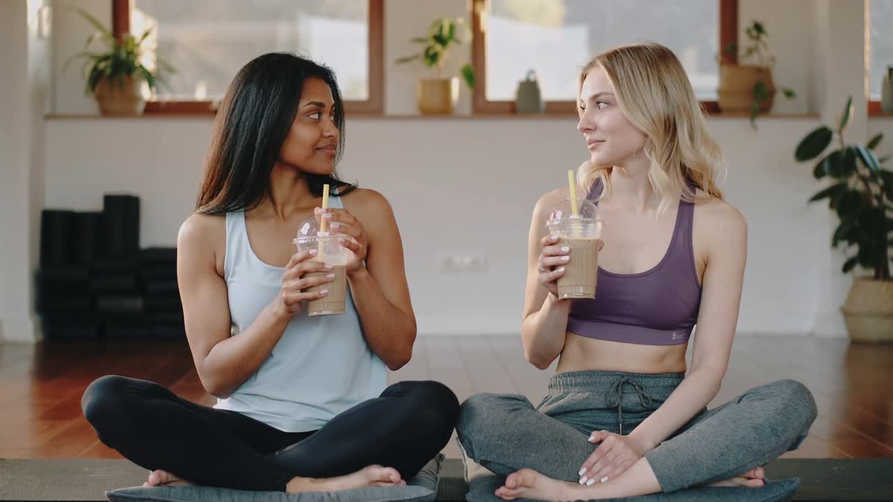 Sipping iced drinks, women in sportswear making eye contact, chatting on mats holding yellow straws