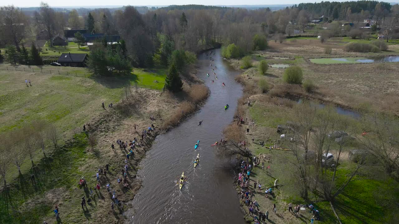 Aerial drone moves backwards above reflected river while many kayakers paddling forward, rowing competition at the Võhandu Marathon surrounded by spring rural landscape. Spectators along the shore