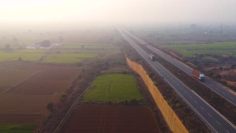 Toma-Aérea-De-Drones-De-Camiones-Moviéndose-Por-Una-Carretera-A-Través-De-Granjas-En-La-Zona-Rural-De-Gwalior-De-La-India