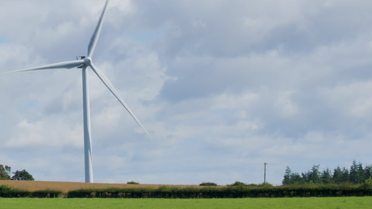 A large wind turbine spins steadily in a rural landscape near Edinburgh, Scotland. Daylight, wide shot, static camera, cloudy sky, green fields