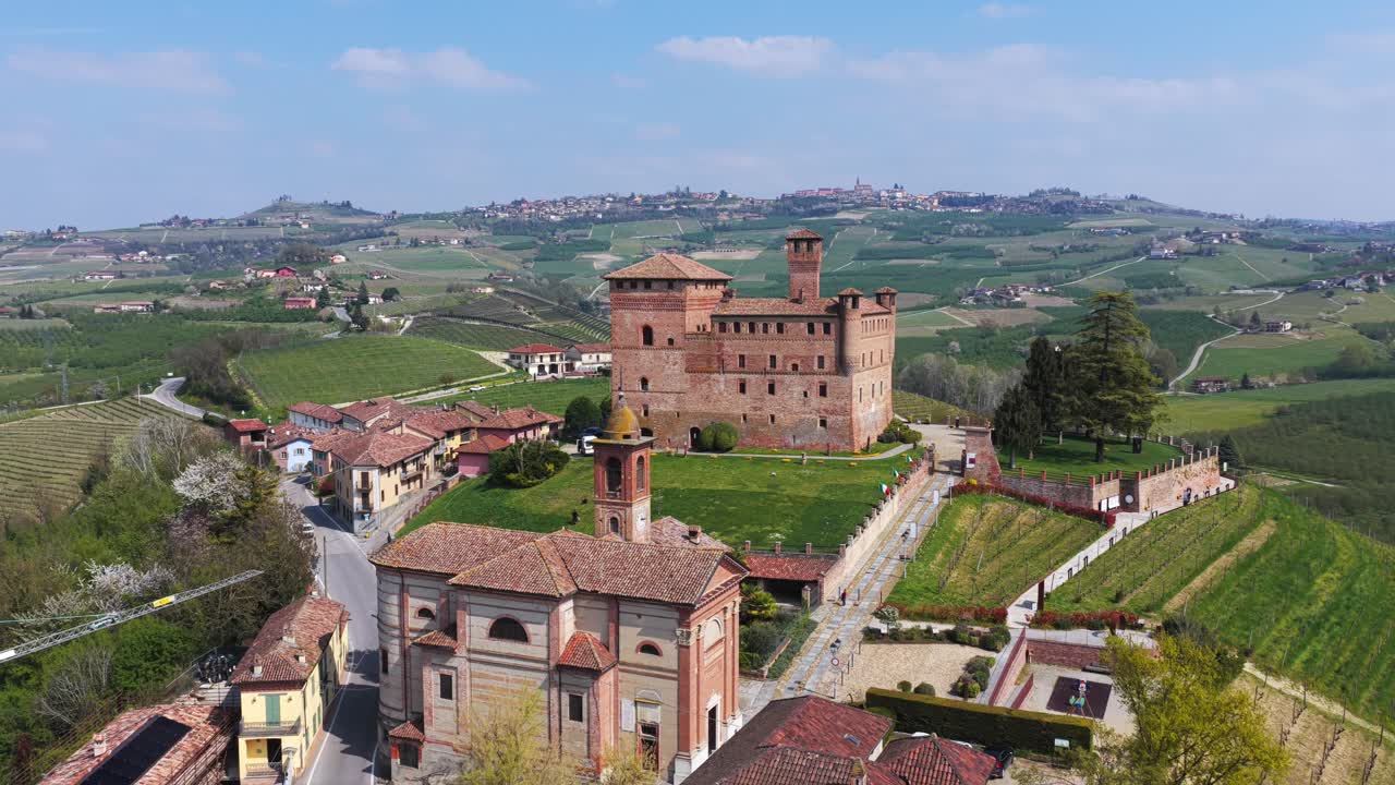 Dolly backward aerial shot of Grinzane Cavour, revealing the majestic brick castle, church tower, and surrounding vineyards of the Langhe region under a bright sky and rolling countryside