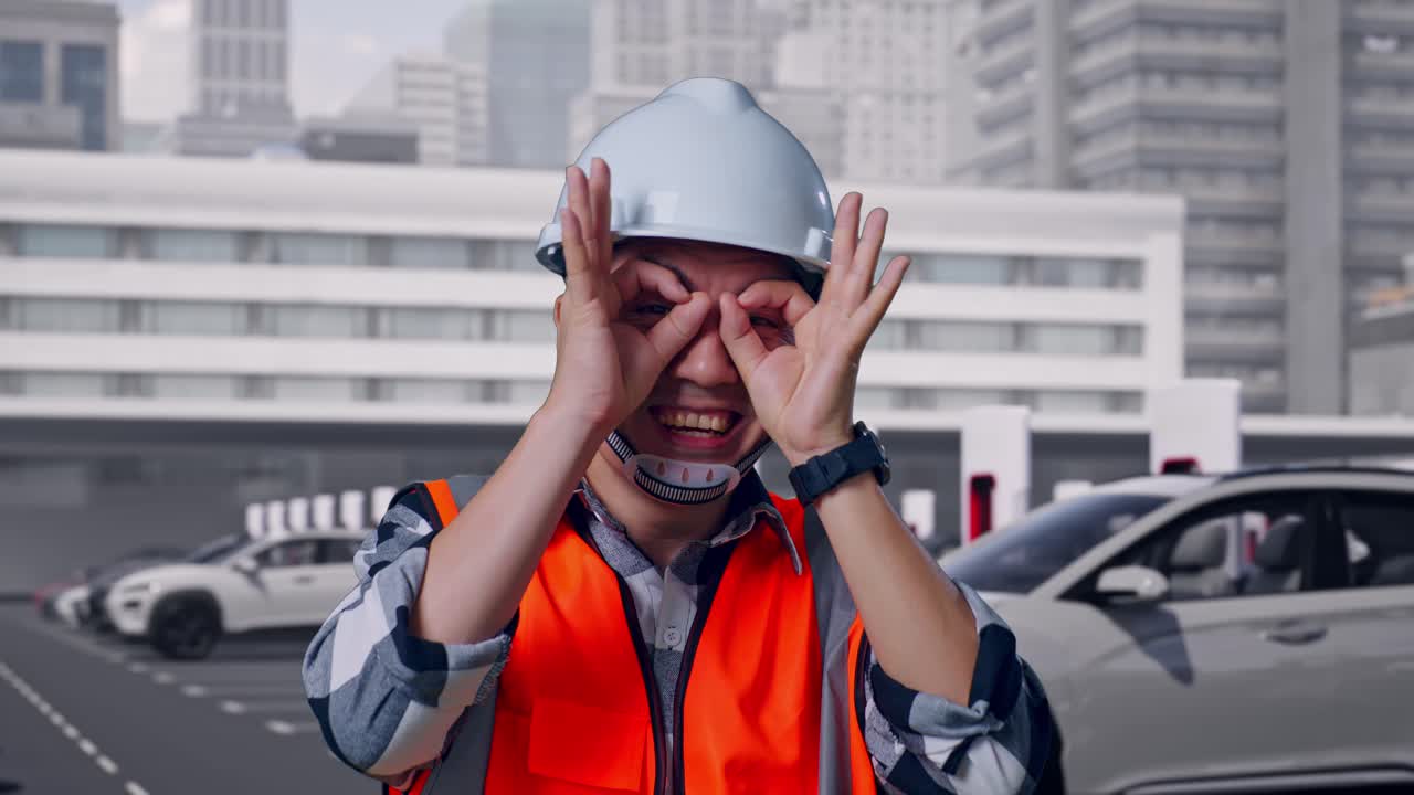 Nærbillede af asiatisk mandlig ingeniør med sikkerhedshjelm, der viser OK-tegn over øjet og smiler til kameraet, mens han står, ved elbiler der oplader på en moderne ladestation i byen.